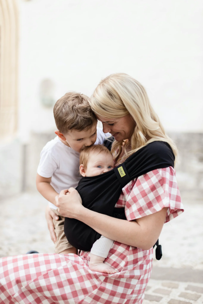 Boba carrier being use by a mom with a baby sitting comfortably. Her toddler son is kissing the baby's head. They look happy.