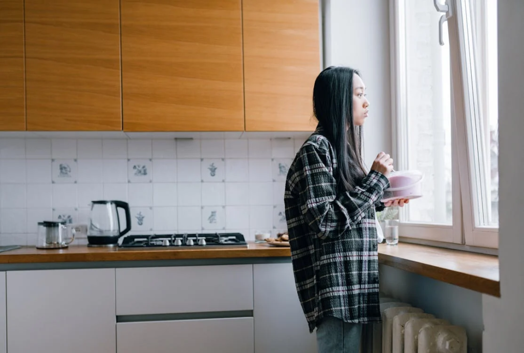 A woman eating a tub of ice cream while looking out the window. She appears sad. Emotional eating.