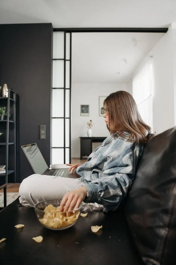 A women eating potato chips while using her laptop. She is sitting on a comfy sofa. Stress eating.